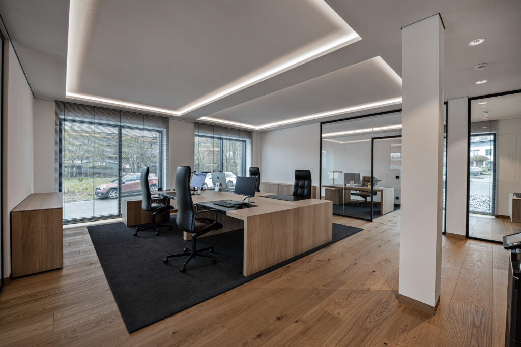 Åpent kontorlandskap med store vindusflater, arbeidsstasjoner og tregulv.